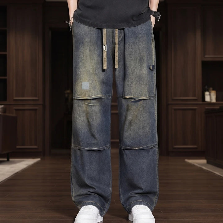 Man wearing distressed, faded blue wide-leg jeans with panel details, a drawstring waist, and white sneakers.