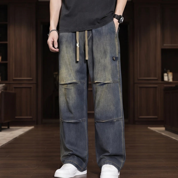 Man wearing loose-fit, faded dark blue denim wide-leg jeans with knee panel details, a drawstring waist, and white sneakers.