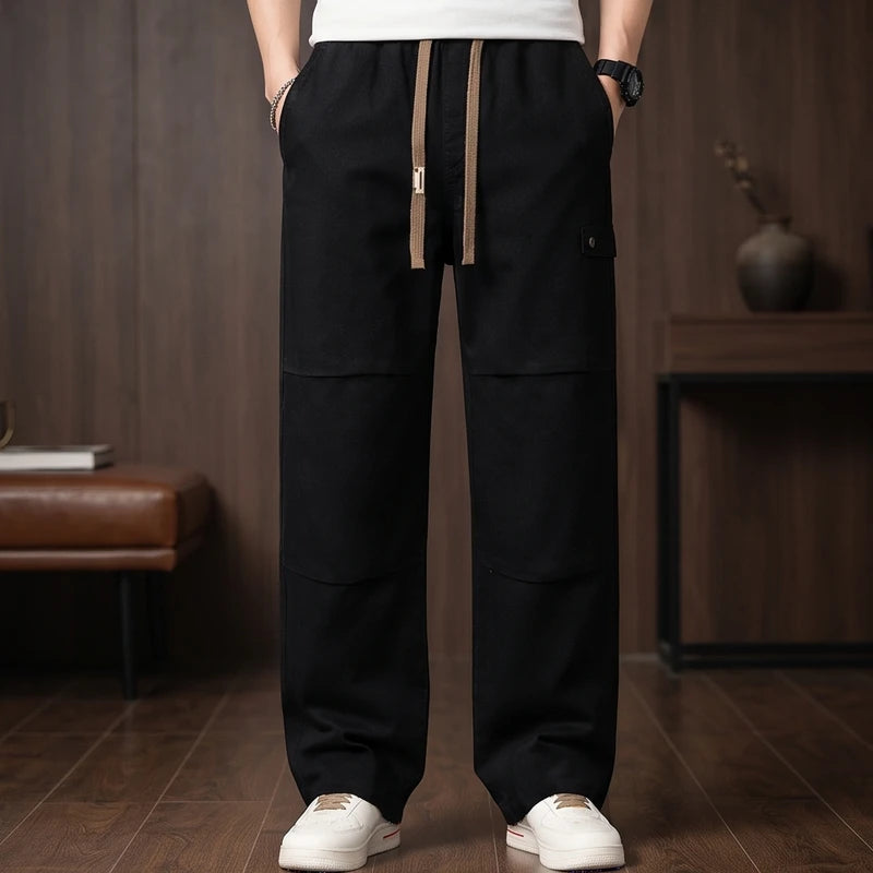 Man wearing black wide-leg drawstring pants with knee panels and a small side pocket, standing on a wooden floor.