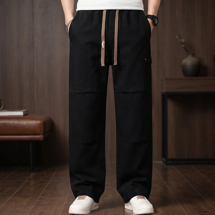 Man wearing black wide-leg drawstring pants with knee panels and a small side pocket, standing on a wooden floor.