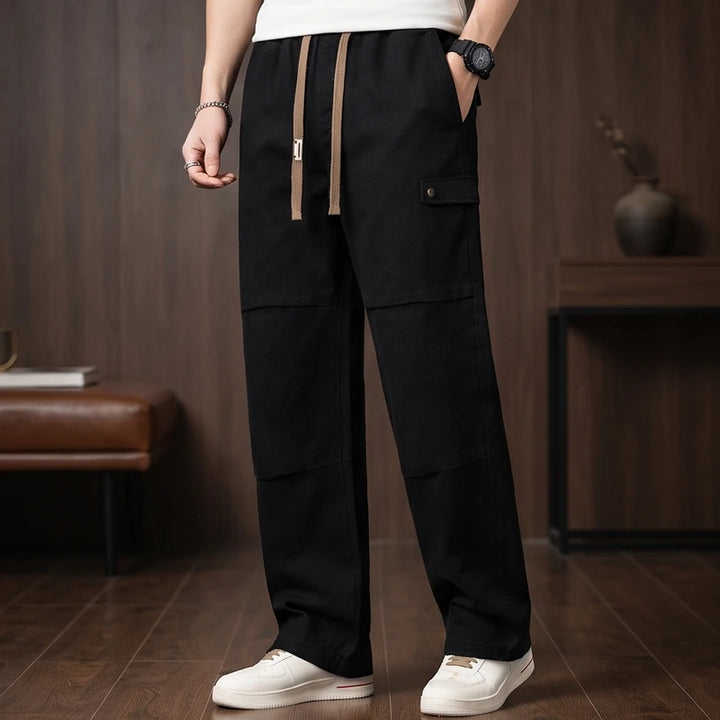 Man wearing black wide-leg cargo pants with a brown drawstring, white sneakers, and a watch, standing indoors.