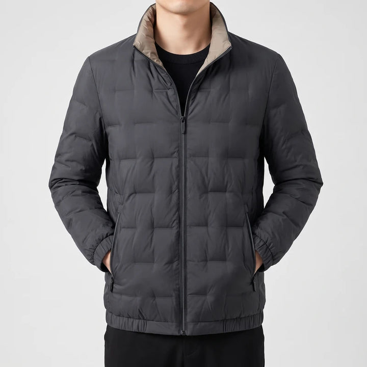 Man wearing a dark gray puffer jacket with a light brown collar and zippered pockets, hands in pockets.
