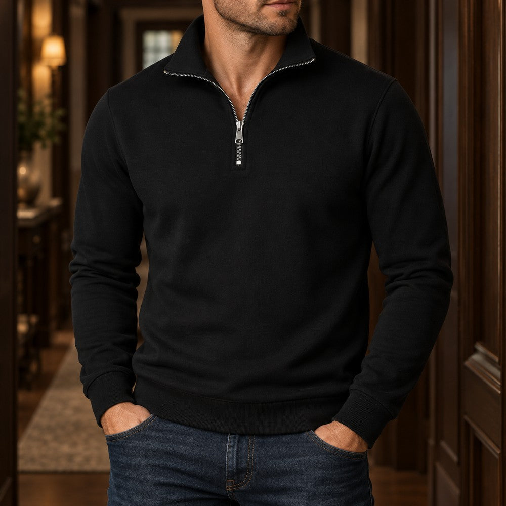 Man wearing a black quarter-zip sweatshirt with a silver zipper, standing with hands in dark blue jeans pockets, indoors.