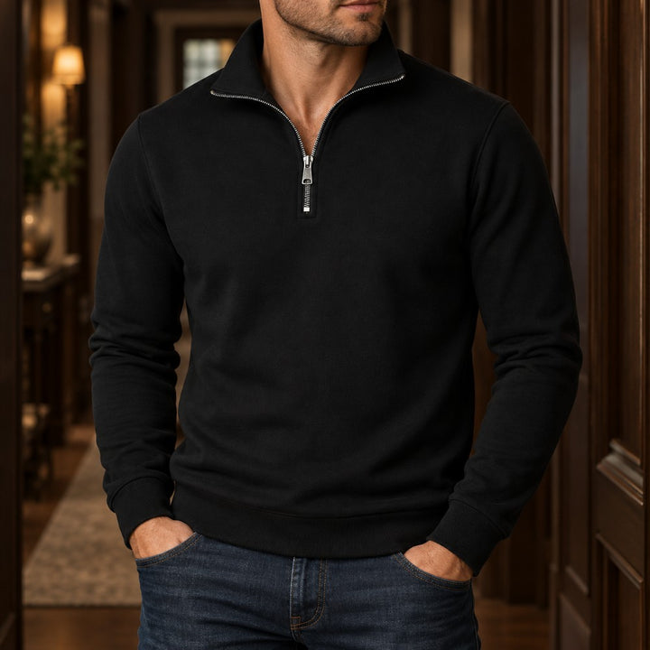 Man wearing a black quarter-zip sweatshirt with a silver zipper, standing with hands in dark blue jeans pockets, indoors.