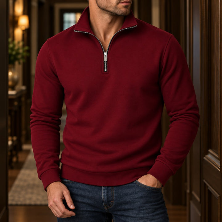 Man wearing a deep red quarter-zip sweatshirt with a silver zipper, blue jeans, standing in a well-lit hallway.