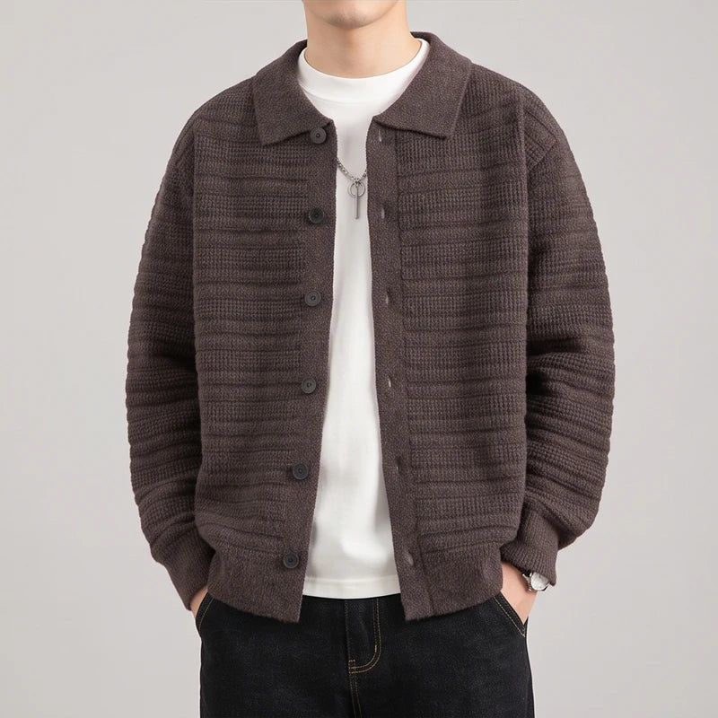Man wearing a brown textured knit button-front cardigan over a white t-shirt and dark jeans, hands in pockets.