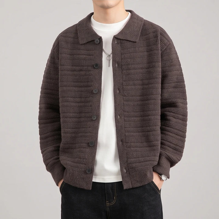 Man wearing a brown textured knit button-front cardigan over a white t-shirt and dark jeans, hands in pockets.