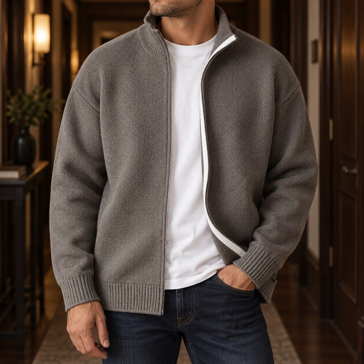 Man wearing an open gray knit cardigan sweater over a white t-shirt, with one hand in his jeans pocket.