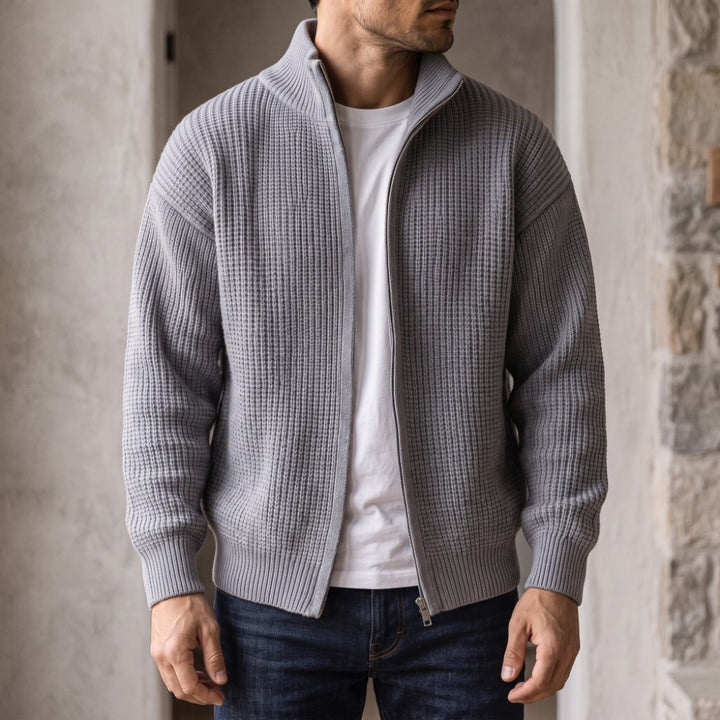 Man wearing an open light gray waffle knit zip-up cardigan over a white t-shirt and blue jeans.