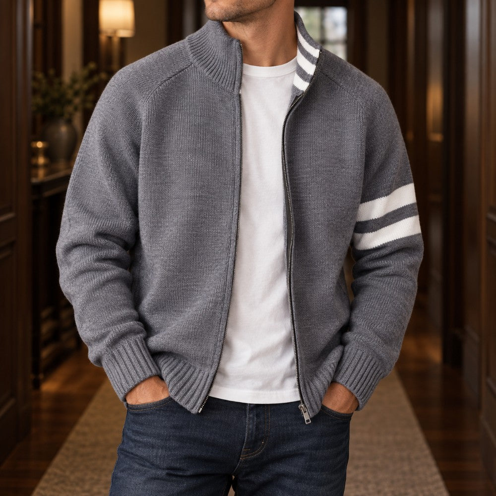Man wearing a grey knit zip-up cardigan with white stripes on the sleeve, over a white t-shirt, standing indoors.