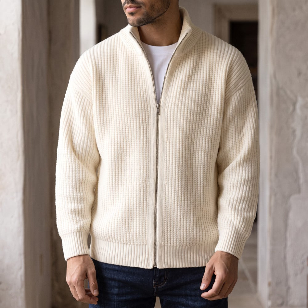 Man wearing a cream-colored, ribbed knit zip-up sweater over a white t-shirt and dark jeans in a hallway.