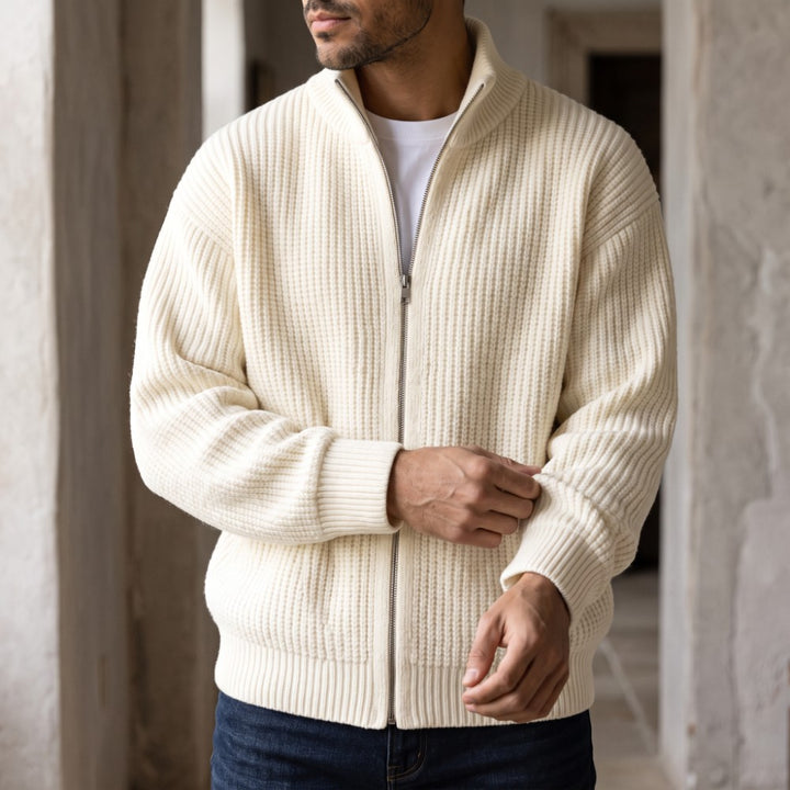 Man wearing a cream waffle-knit cardigan with a silver zipper, white t-shirt, and dark jeans, rolling up his sleeve.
