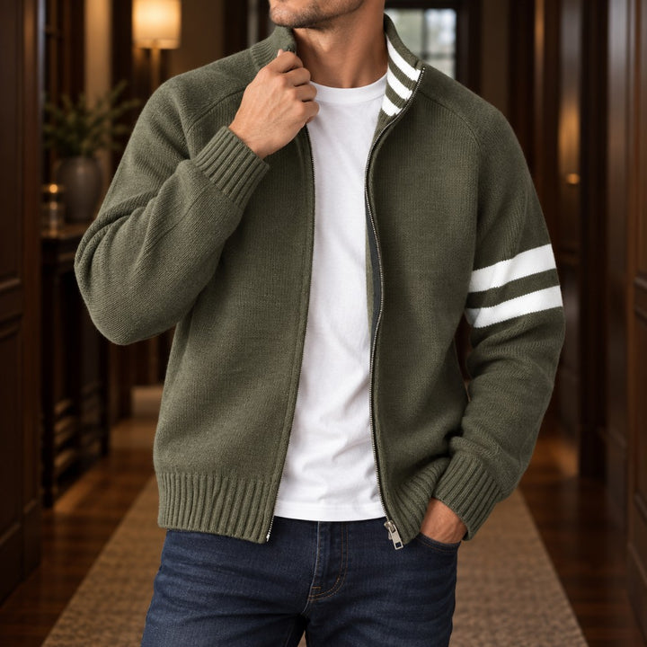 Man wearing an unzipped olive green knit cardigan with white sleeve stripes over a white tee, in a hallway.