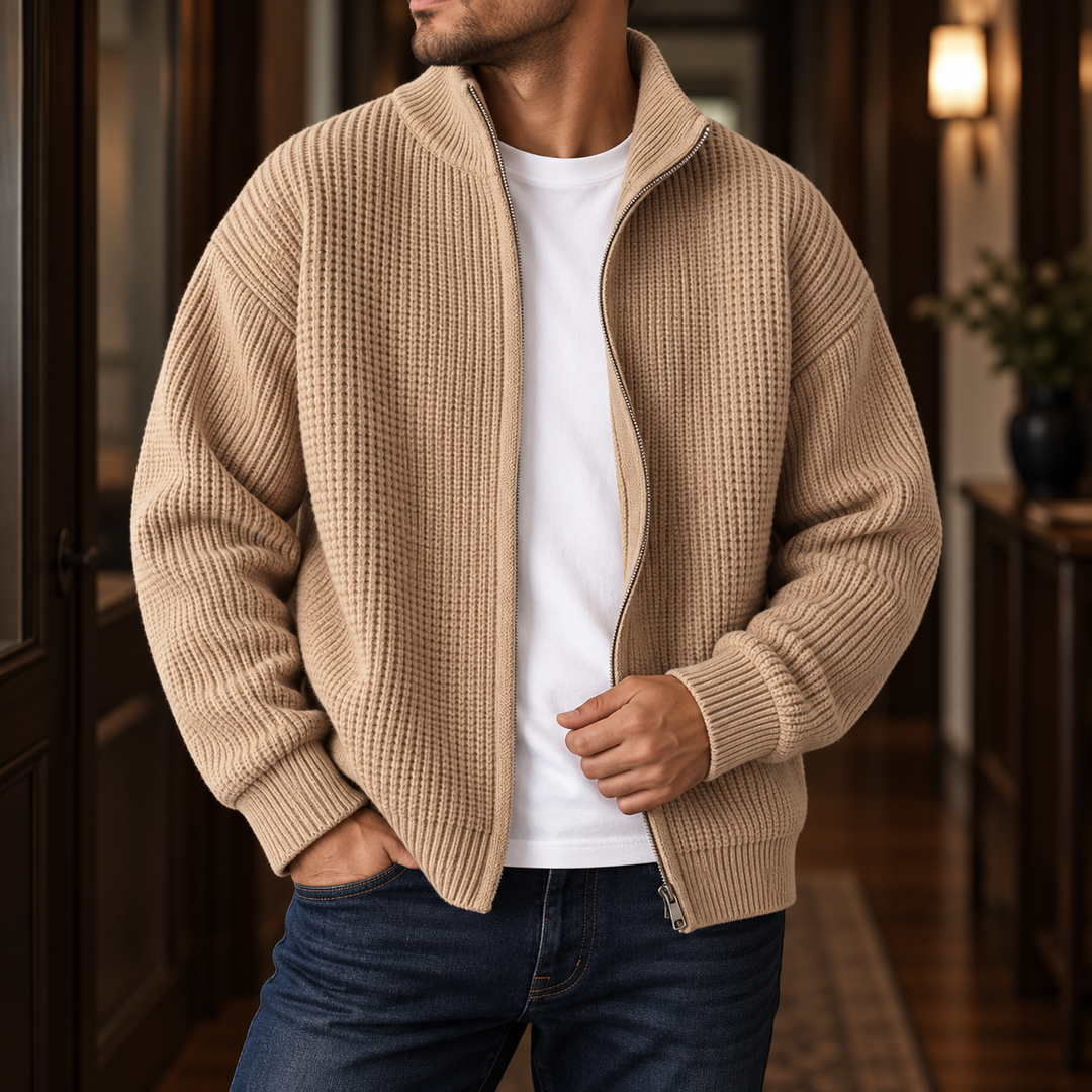 Man wearing a beige waffle-knit zippered sweater with a white t-shirt and blue jeans, standing indoors.