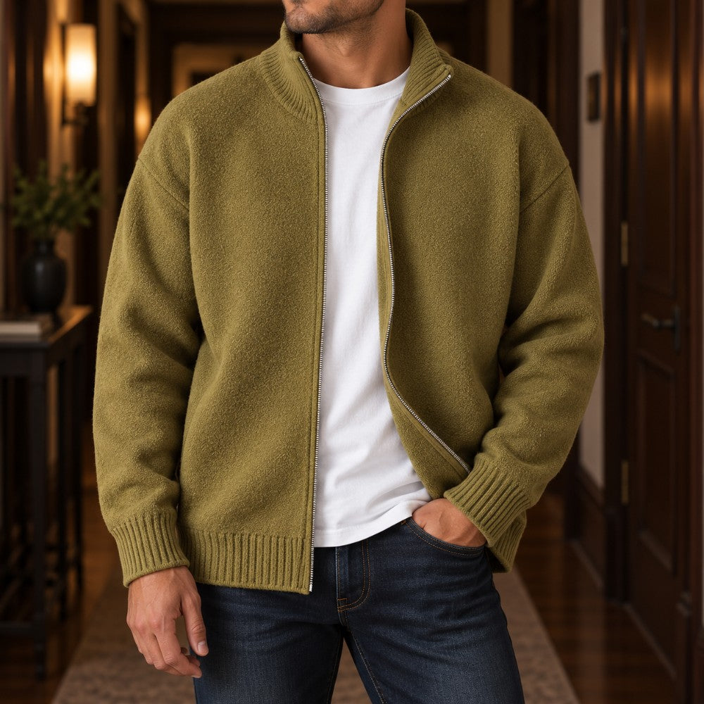 Man wearing a fuzzy olive green knit cardigan with a silver zipper over a white t-shirt and dark jeans in a hallway.