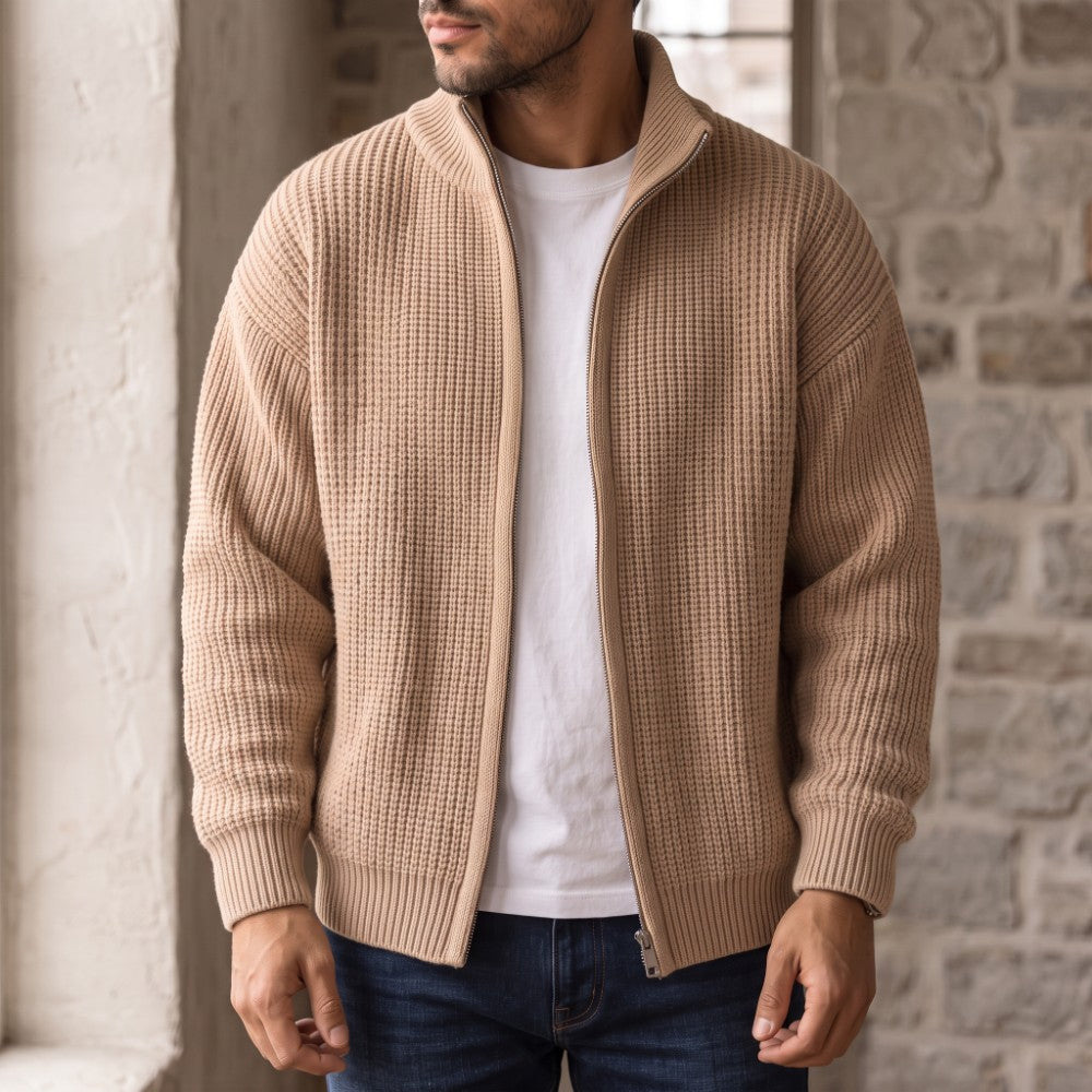 Man wearing a light brown waffle-knit full-zip cardigan over a white t-shirt and blue jeans, standing indoors.