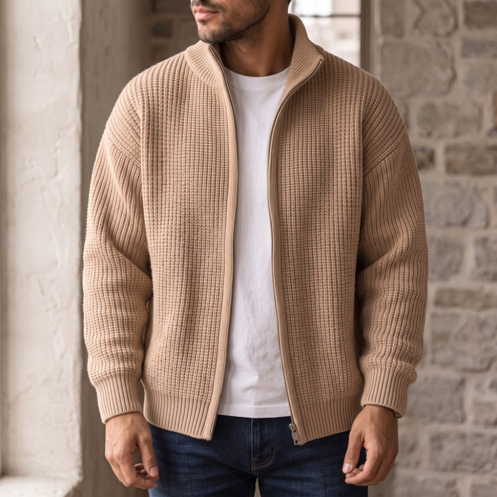 Man wearing a light brown waffle-knit full-zip cardigan over a white t-shirt and blue jeans, standing indoors.