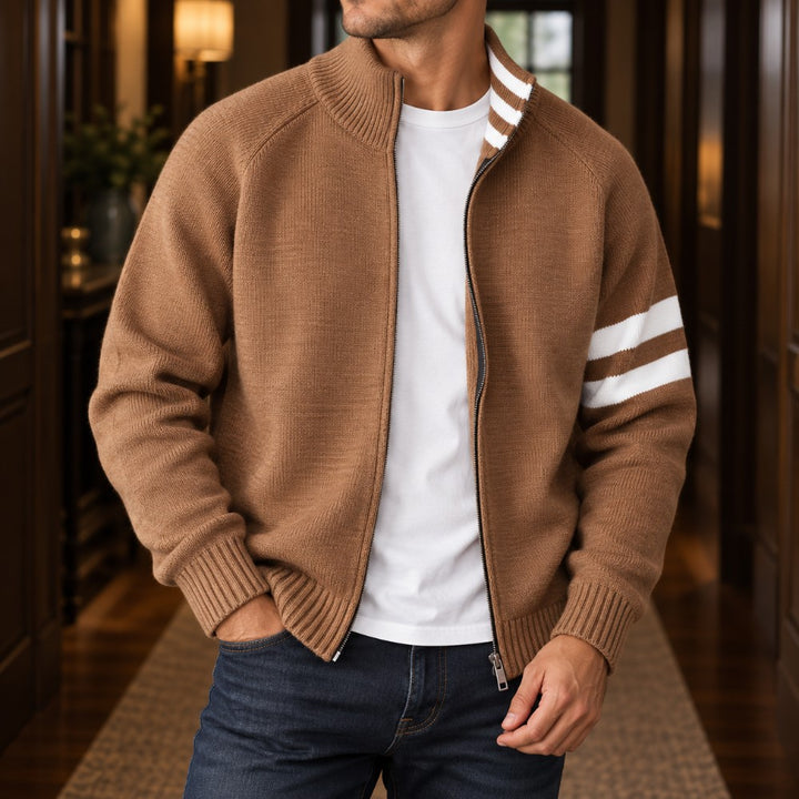 Man wearing a brown knit zippered cardigan with white sleeve stripes, a white t-shirt, and dark blue jeans.