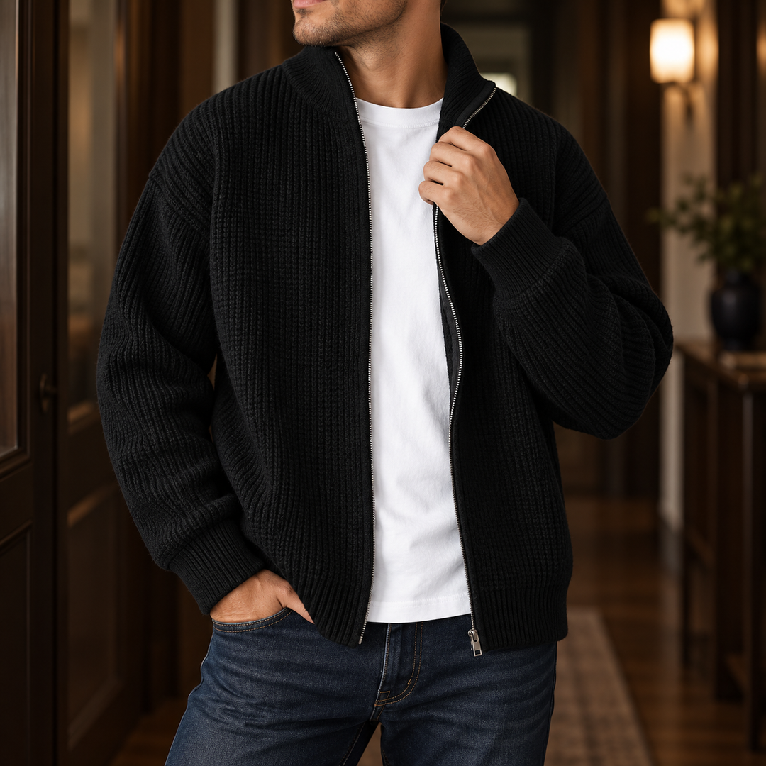 Man wearing a black ribbed knit full-zip cardigan sweater over a white t-shirt and dark blue jeans indoors.