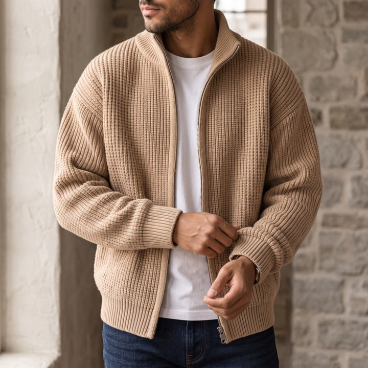 Man wearing an open tan waffle knit cardigan over a white t-shirt and dark jeans, adjusting the sleeve.