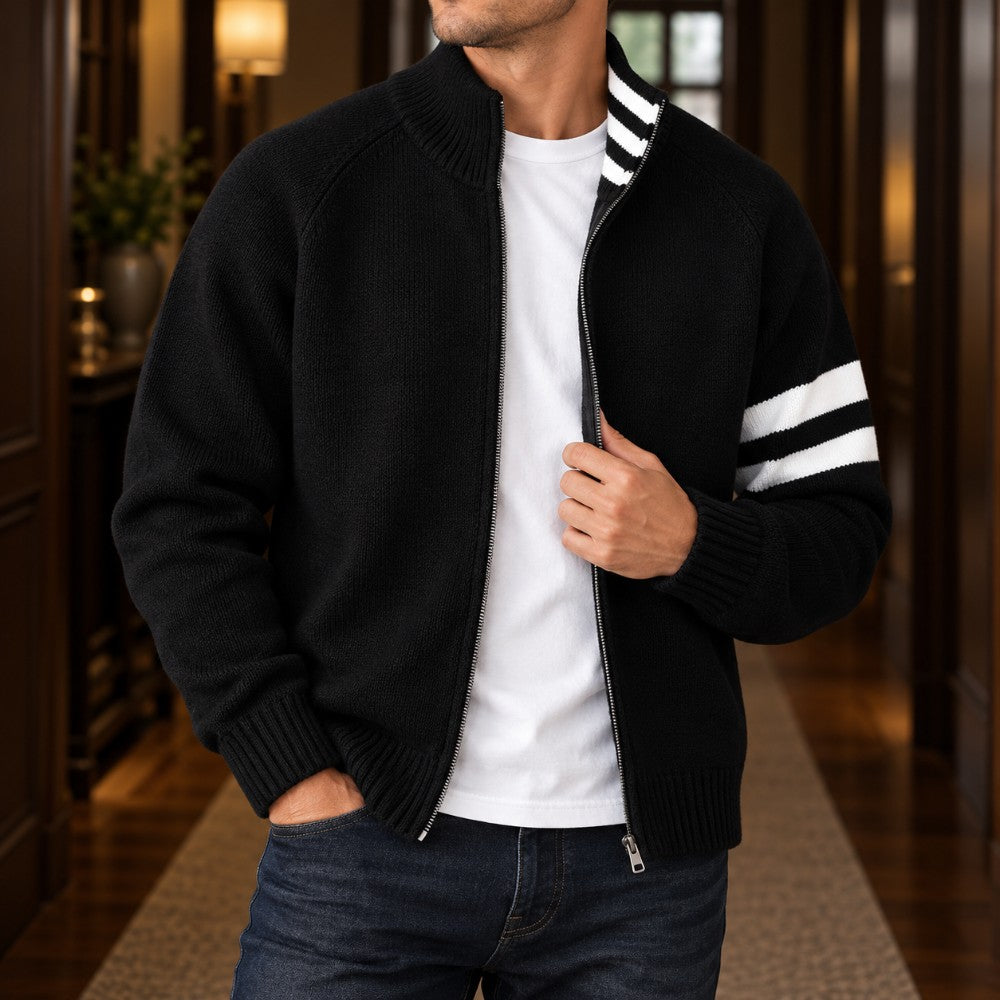 Man wearing a black knit zip-up cardigan with white stripes on the sleeve over a white t-shirt, standing indoors.