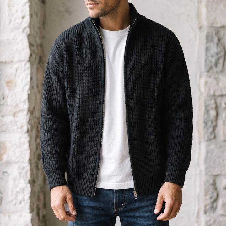 Man wearing an unzipped black waffle knit sweater over a white t-shirt and blue jeans, standing against a stone wall.