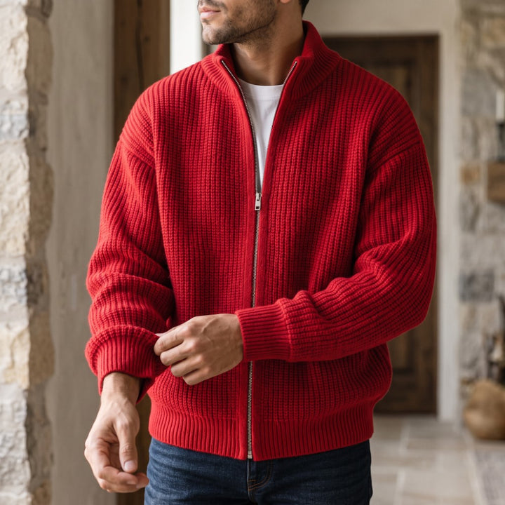 Man adjusting the cuff of a chunky red knit cardigan with a silver zipper, worn over a white shirt and blue jeans.