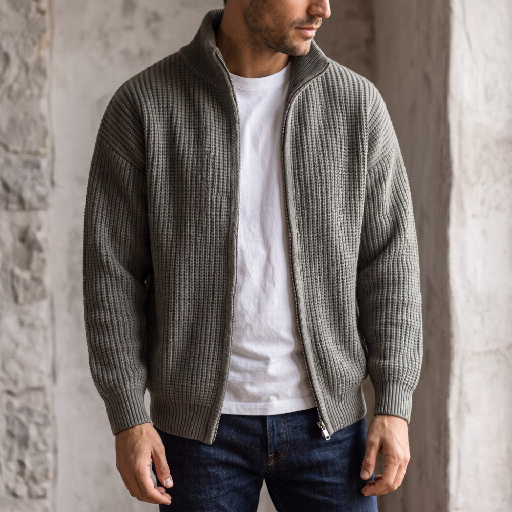 Man wearing an unzipped gray waffle-knit sweater over a white t-shirt and dark blue jeans against a stone wall.