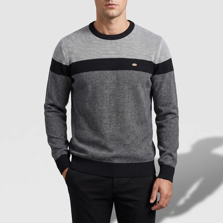 Man wearing a crew neck colorblock knit sweater with gray melange body and a black horizontal chest stripe, standing.