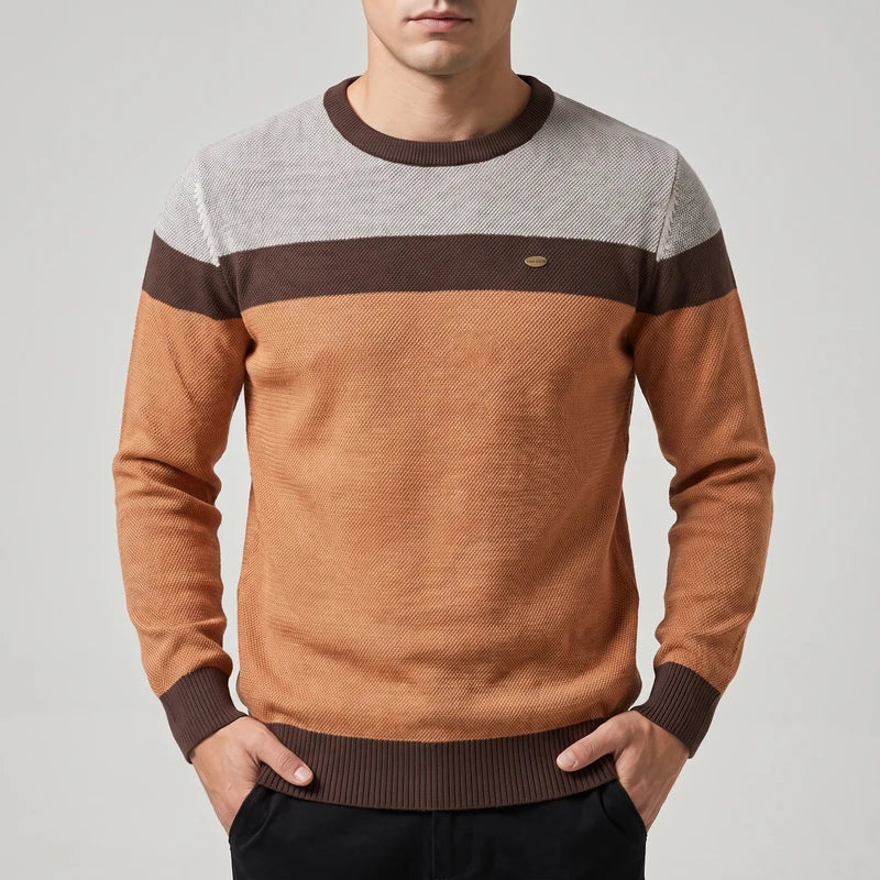 Man wearing a knit sweater with light gray, brown, and orange color blocks, hands in black pants pockets.