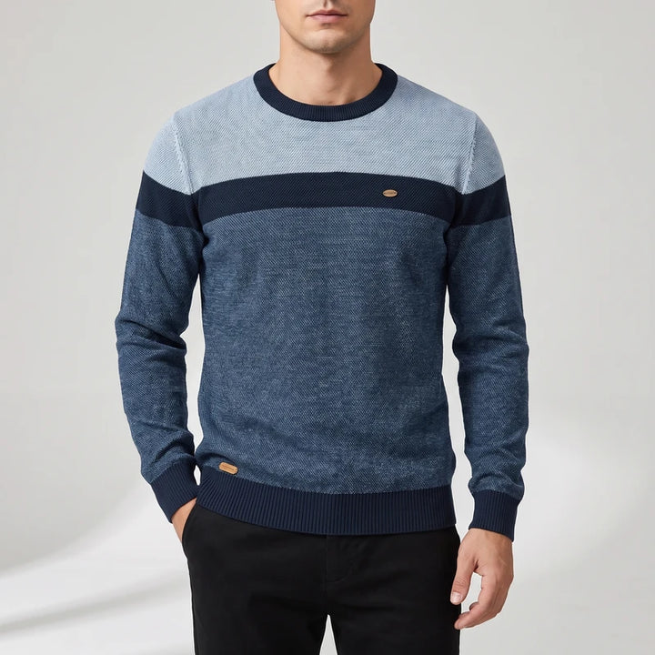Man wearing a crew neck colorblock sweater with light blue, navy, and dark blue knit textures and a small brown tag.
