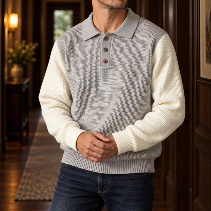 Man wearing a light gray knit polo with cream long sleeves and brown buttons, standing in a wood-paneled hallway.