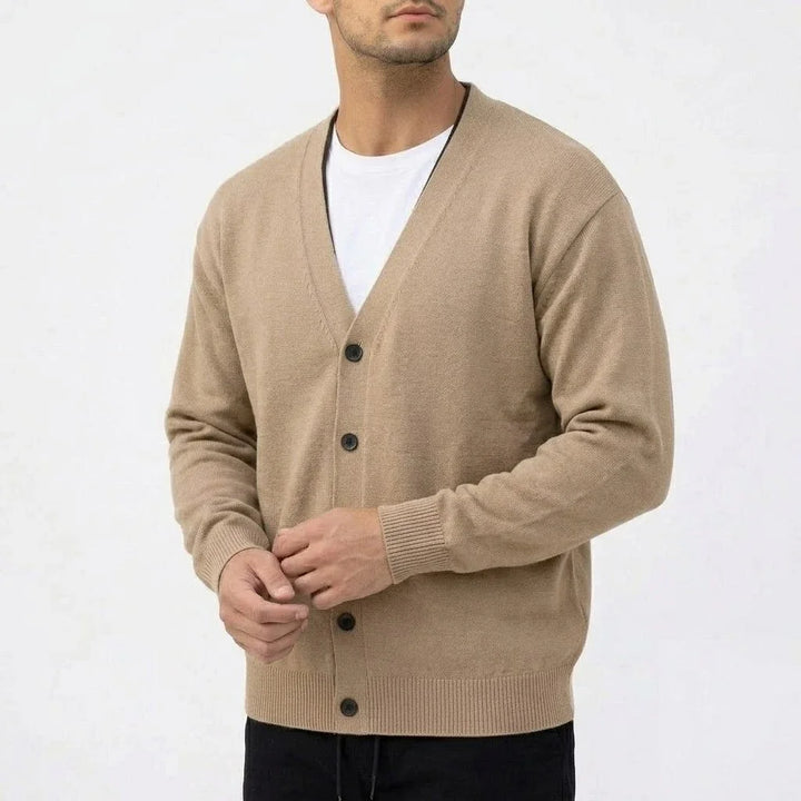 Man wearing a light brown knit V-neck cardigan with dark buttons over a white t-shirt, standing against a white background.
