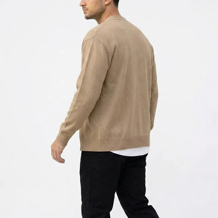 Man wearing a light brown knit cardigan over a white shirt and black pants, shown from the back, walking left.