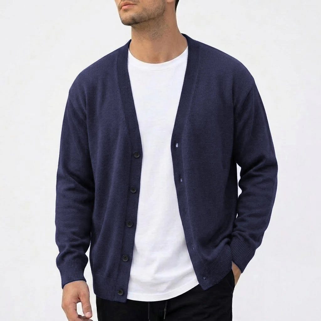 Man wearing an open dark navy V-neck knit cardigan over a white t-shirt, with one hand in his black pant pocket.
