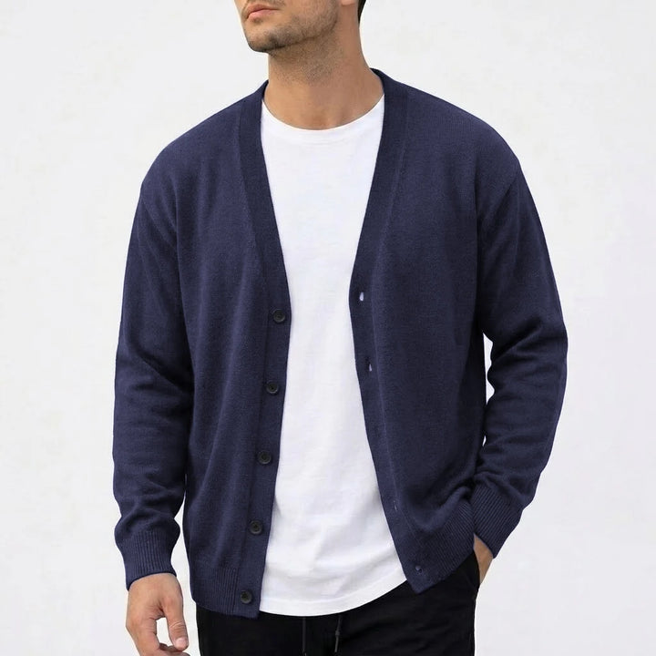 Man wearing an open dark navy V-neck knit cardigan over a white t-shirt, with one hand in his black pant pocket.