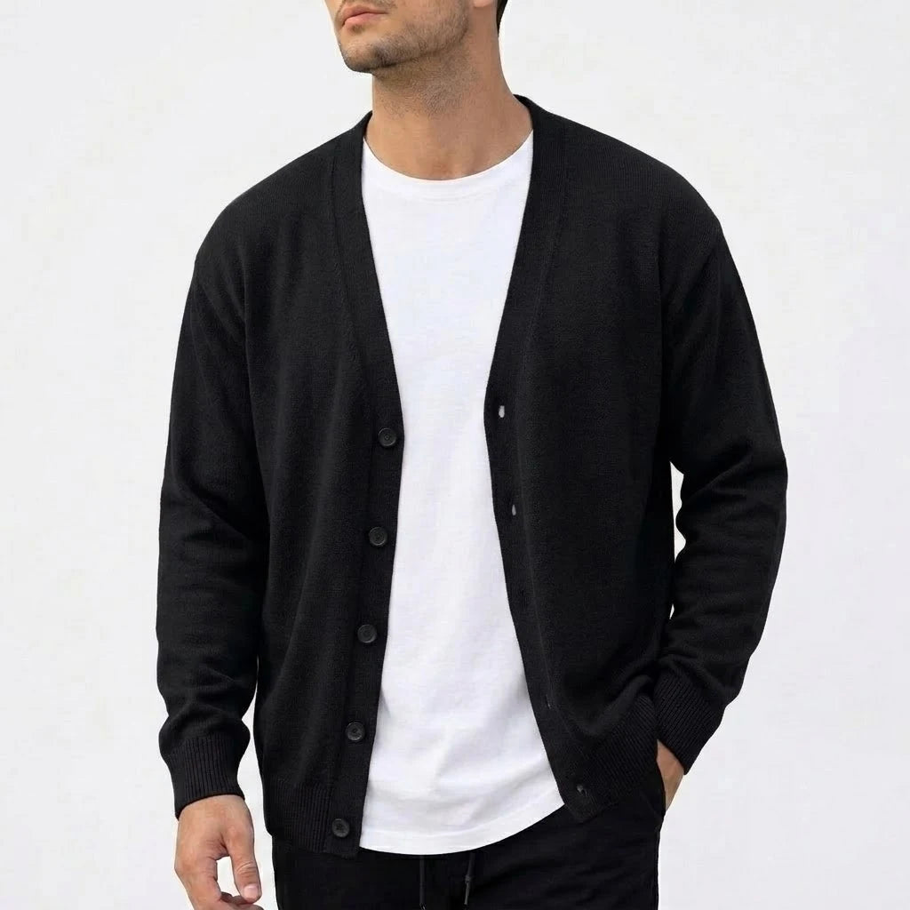 Man wearing a black knit V-neck cardigan over a white t-shirt, hand in pocket against a white background.