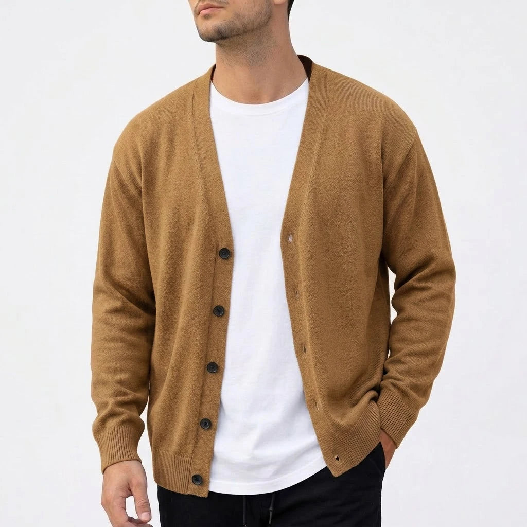 Man wearing an open V-neck cardigan in a warm brown knit over a white t-shirt, with one hand in his black pant pocket.