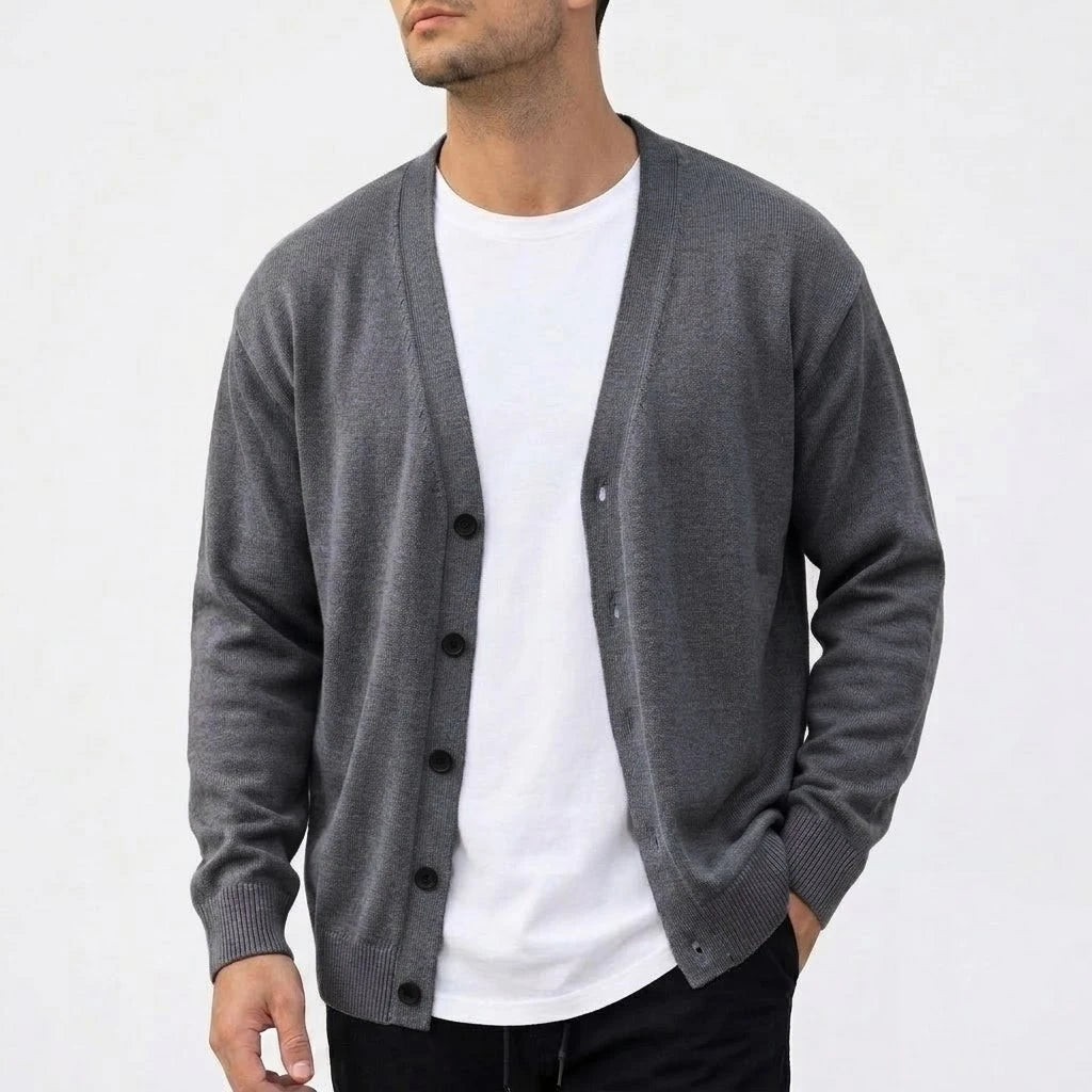 Man wearing a dark gray knit V-neck cardigan with black buttons, over a white t-shirt, standing against a white wall.