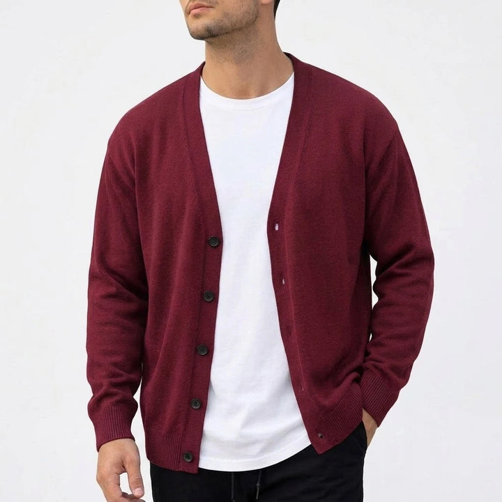 Man wearing a burgundy knit V-neck cardigan with dark buttons open over a white t-shirt, standing against a white background.