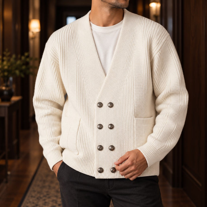 Man wearing a cream ribbed knit double-breasted cardigan with eight dark buttons and two front pockets, standing in a hallway.