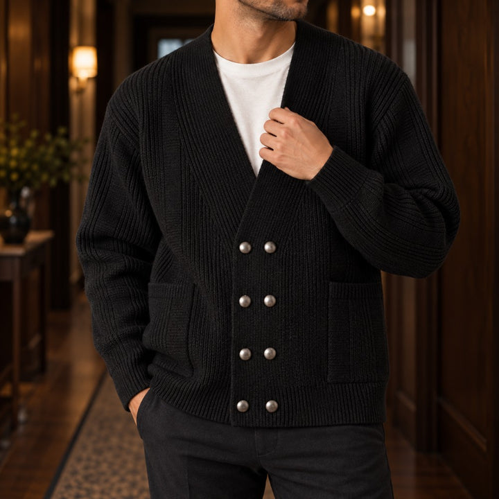 Man wearing a black ribbed double-breasted cardigan with silver buttons and pockets over a white tee, standing indoors.
