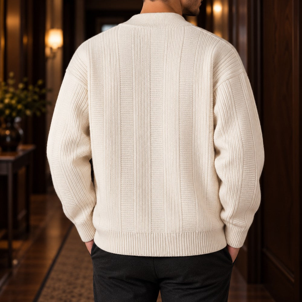 Man wearing an off-white ribbed knit sweater, seen from the back, hands in dark trousers pockets, in a softly lit hallway.