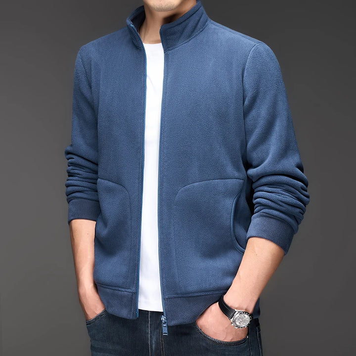 Man wearing a blue fleece zip-up jacket over a white t-shirt, with hands in pockets and a watch on his left wrist.