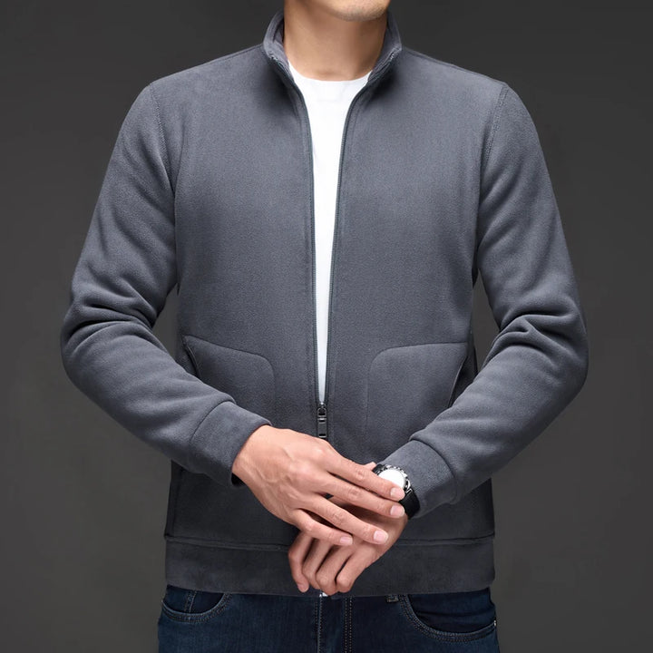 Man wearing a dark gray fleece zip-up jacket over a white t-shirt, with hands clasped in front.