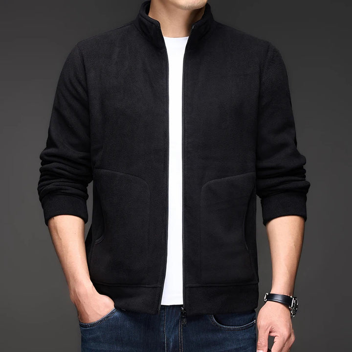 Man wearing a black fleece zip-up jacket over a white t-shirt, standing with hands in blue jeans pockets.