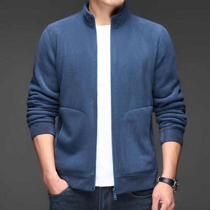 Man wearing an unzipped blue fleece zip-up jacket over a white t-shirt, with hands in dark jeans pockets.