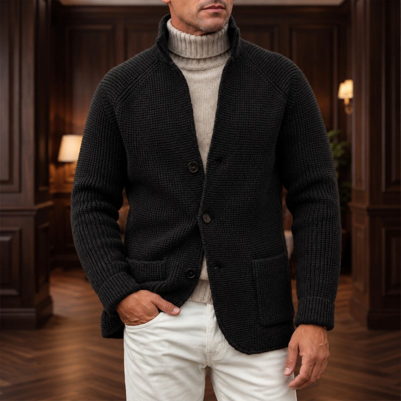 A man wearing a dark charcoal textured knit cardigan over a beige turtleneck sweater and white pants, standing in a dimly lit wood-paneled room.
