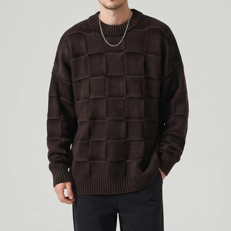 Man wearing an oversized dark brown knit sweater with a raised checkerboard pattern, black pants, and a silver chain.
