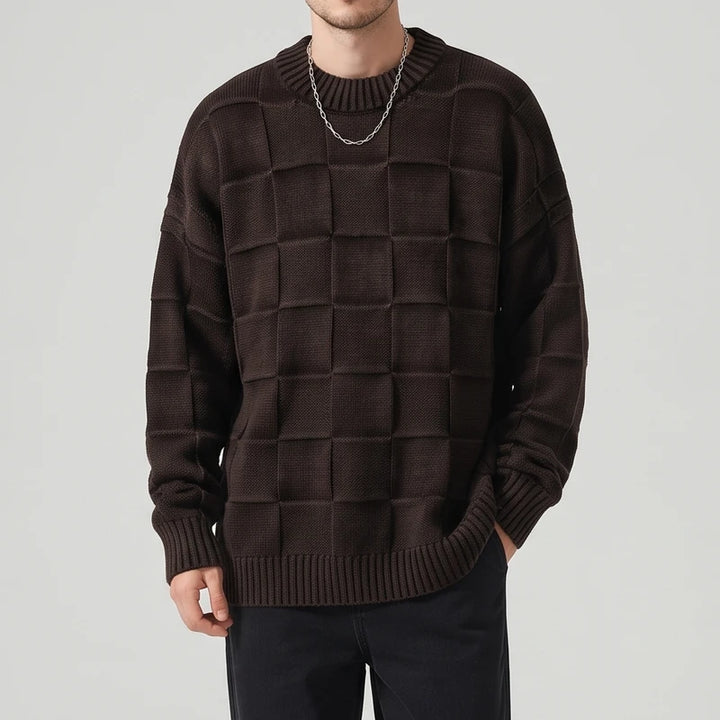 Man wearing an oversized dark brown knit sweater with a raised checkerboard pattern, black pants, and a silver chain.